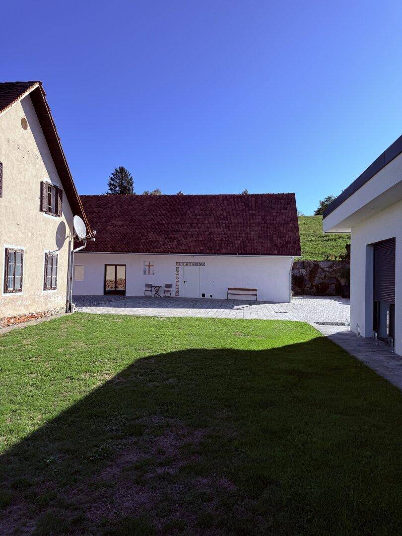 Sonniger Gartenbereich mit Rasenfläche, gepflastertem Weg und zwei Gebäuden im Hintergrund.