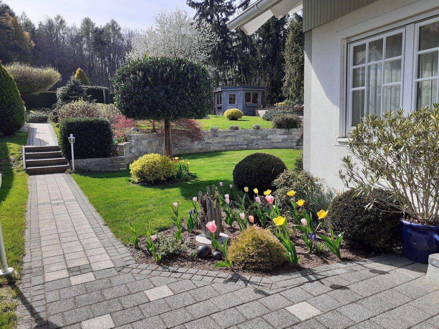 Sonniger Gartenweg mit blühenden Blumenbeeten und gepflegtem Rasenbereich.