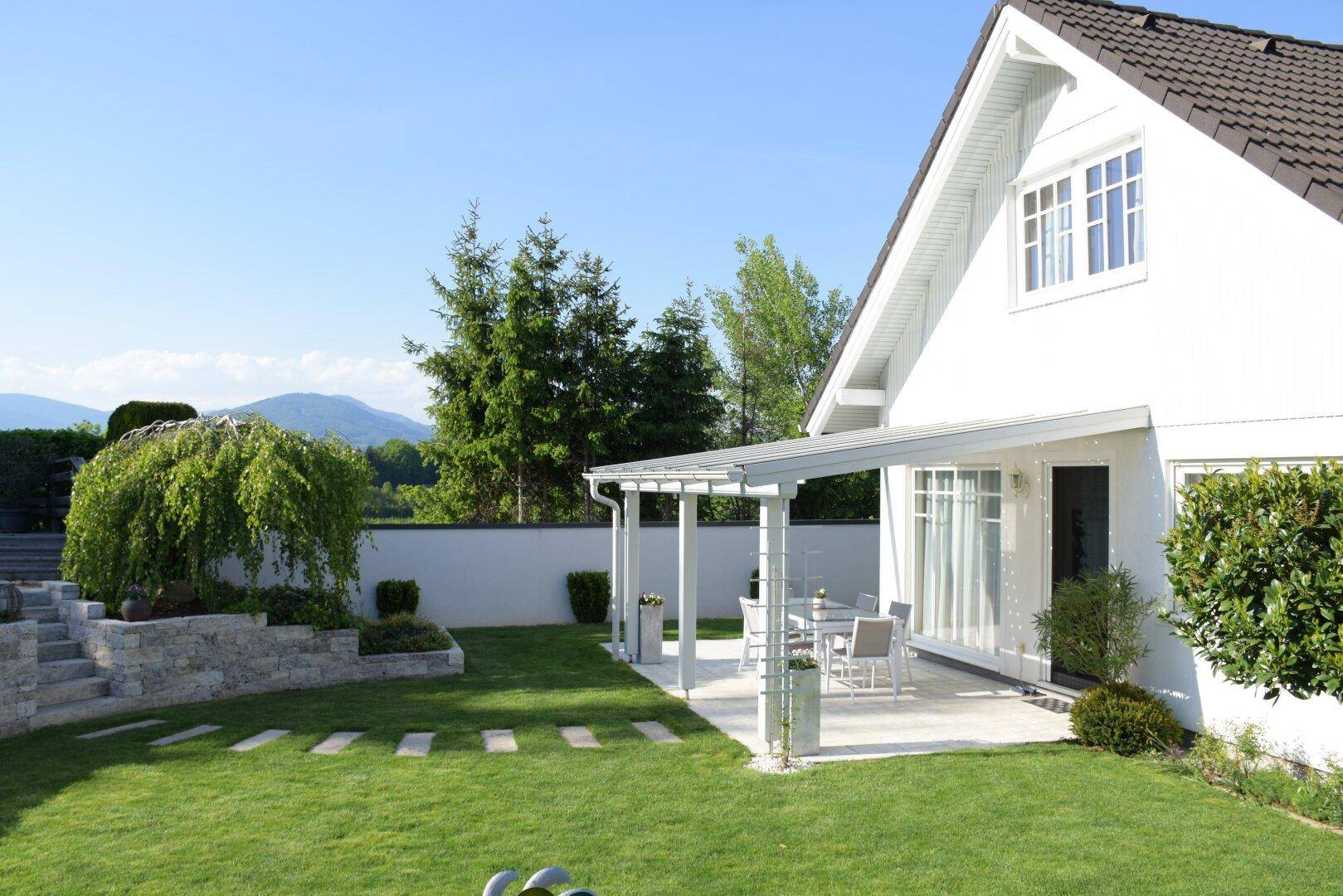 Weißes Haus mit Terrasse und großem Garten, umgeben von grünen Hügeln und Bergen.