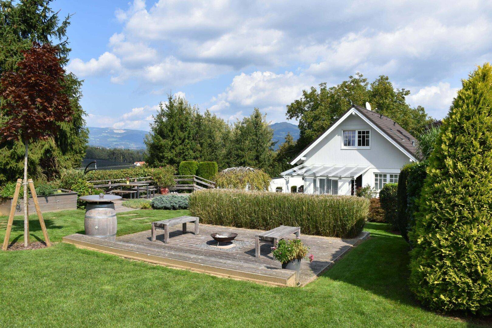 Weitläufiger Garten mit Holzterrasse, Feuerstelle und Sitzbänken im Freien.