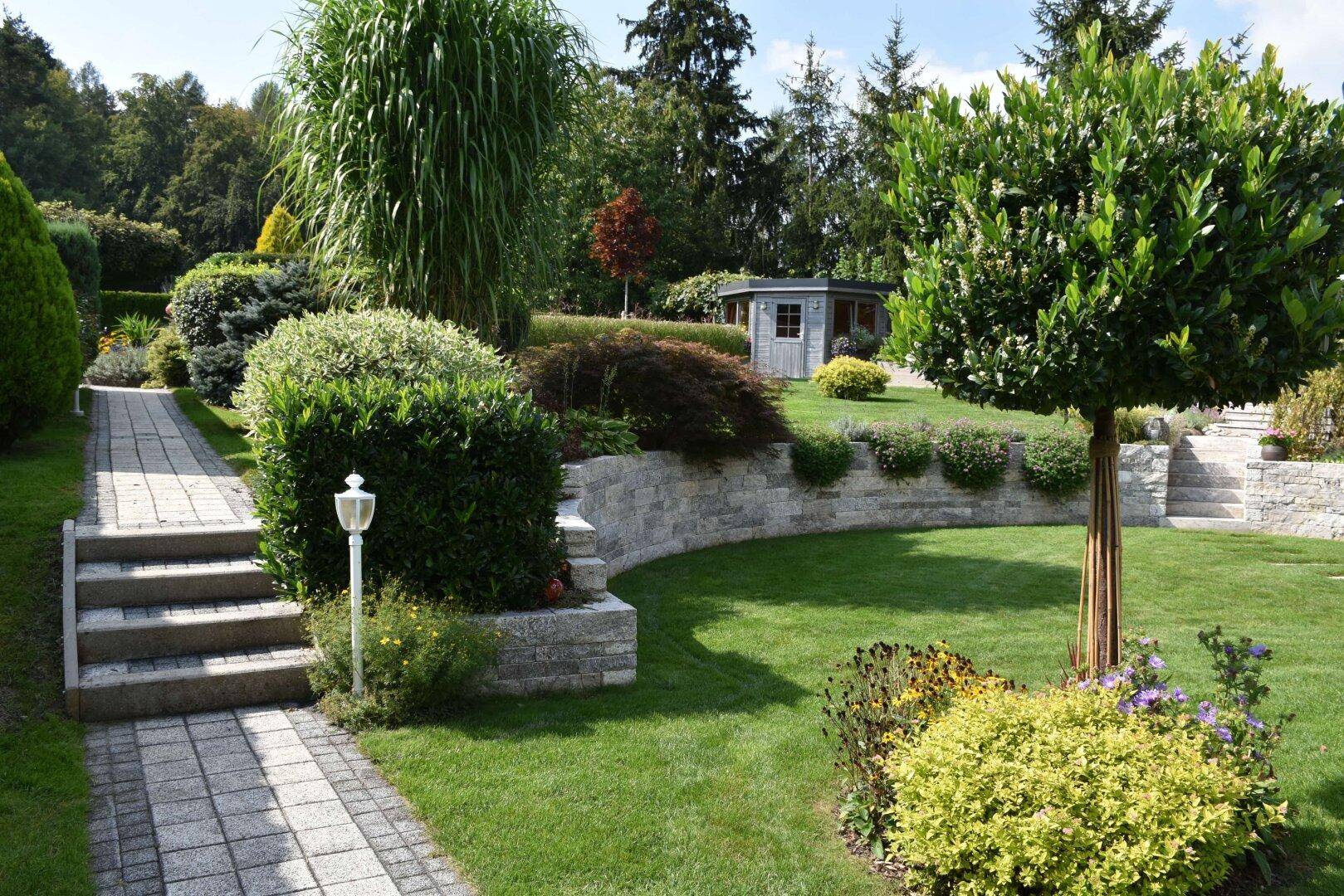 Weitläufiger Garten mit geschwungenen Steinmauern, Rasenflächen und einem Gartenhaus.