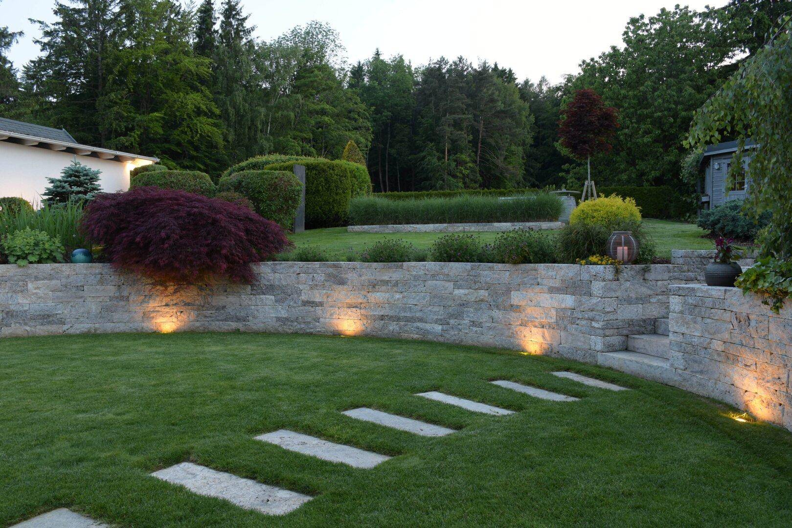 Beleuchteter Gartenbereich am Abend mit Steinmauern, Rasen und Trittplatten.