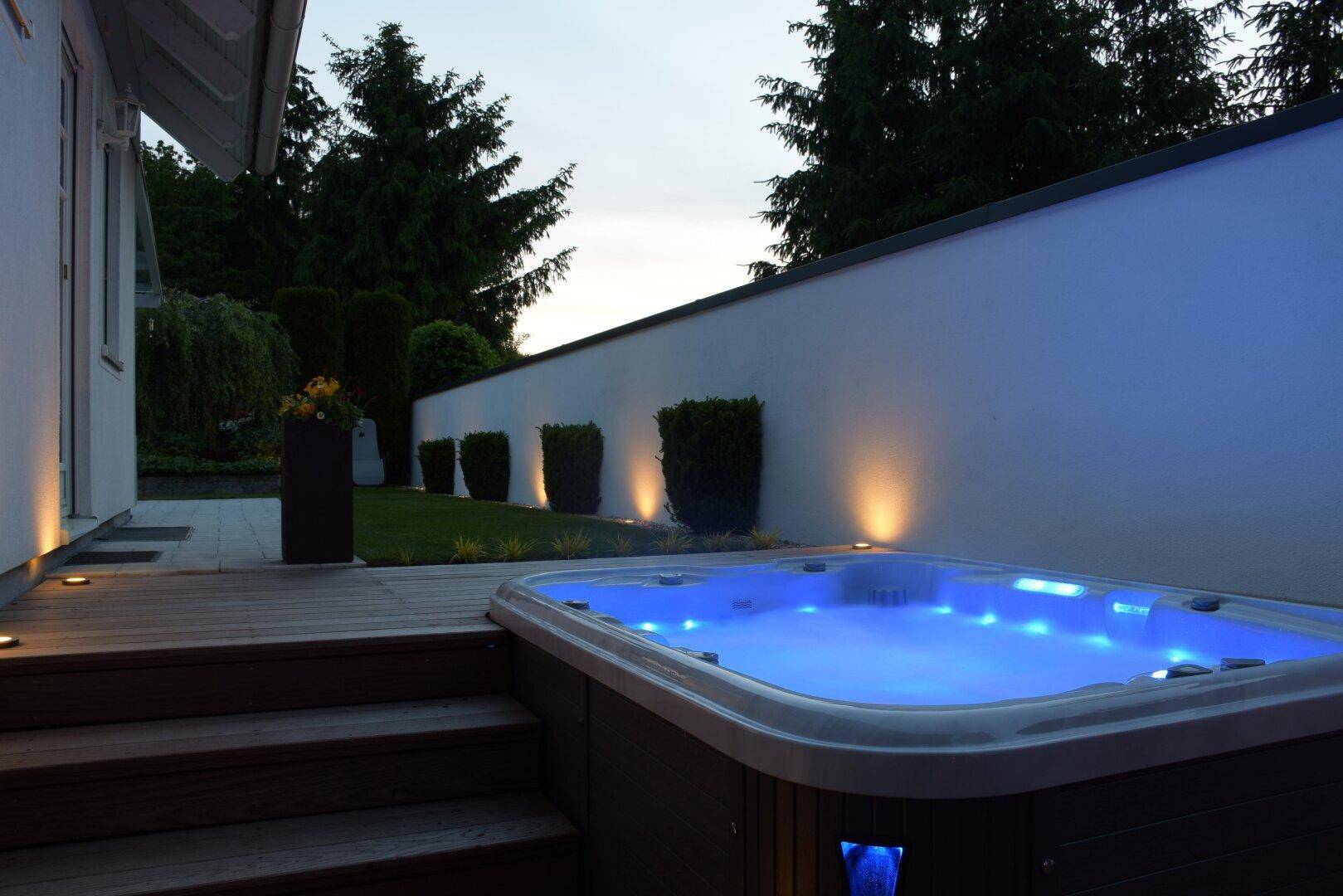 Beleuchteter Whirlpool auf einer Holzterrasse im Garten bei Dämmerung, umgeben von einer weißen Mauer.