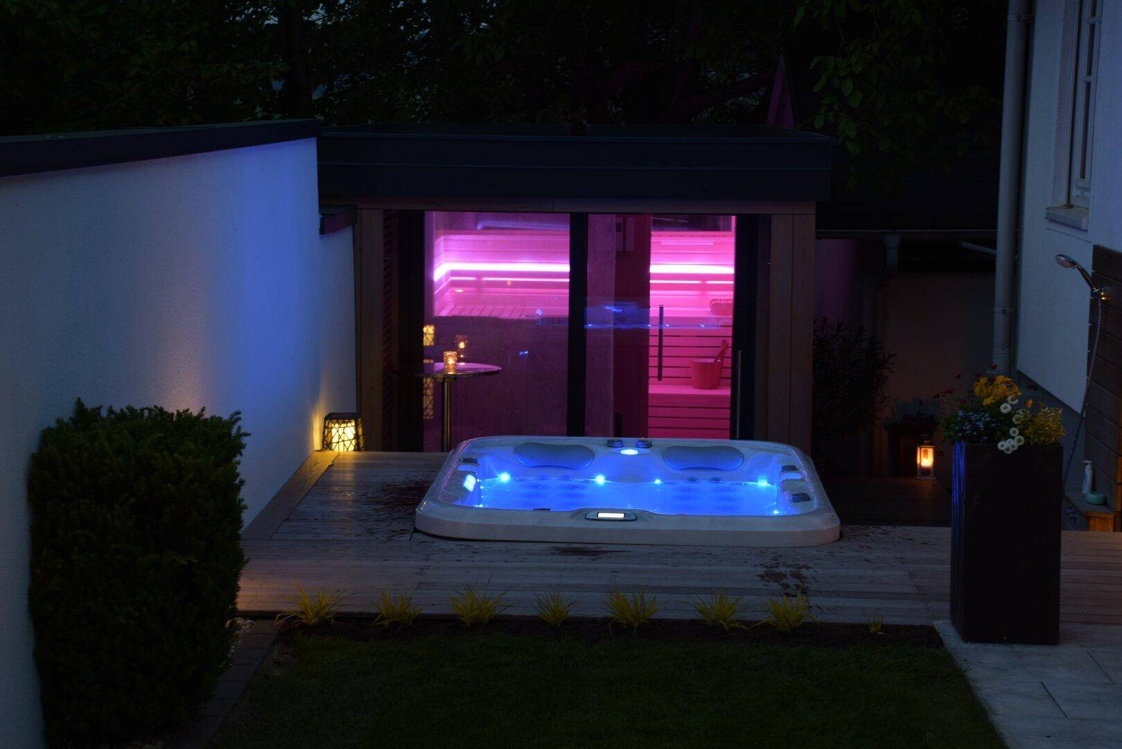 Whirlpool und Sauna mit farbiger Beleuchtung auf einer Holzterrasse im Garten bei Nacht.