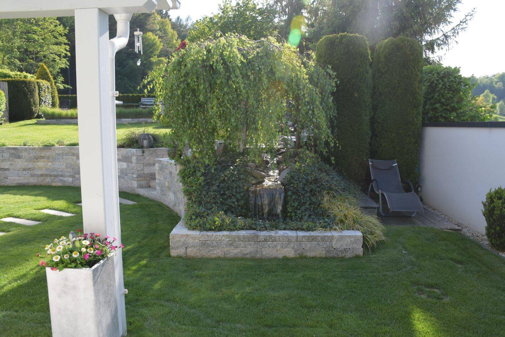 Idyllischer Gartenbereich mit einem kleinen Wasserfall, üppiger Bepflanzung und Liegestuhl.