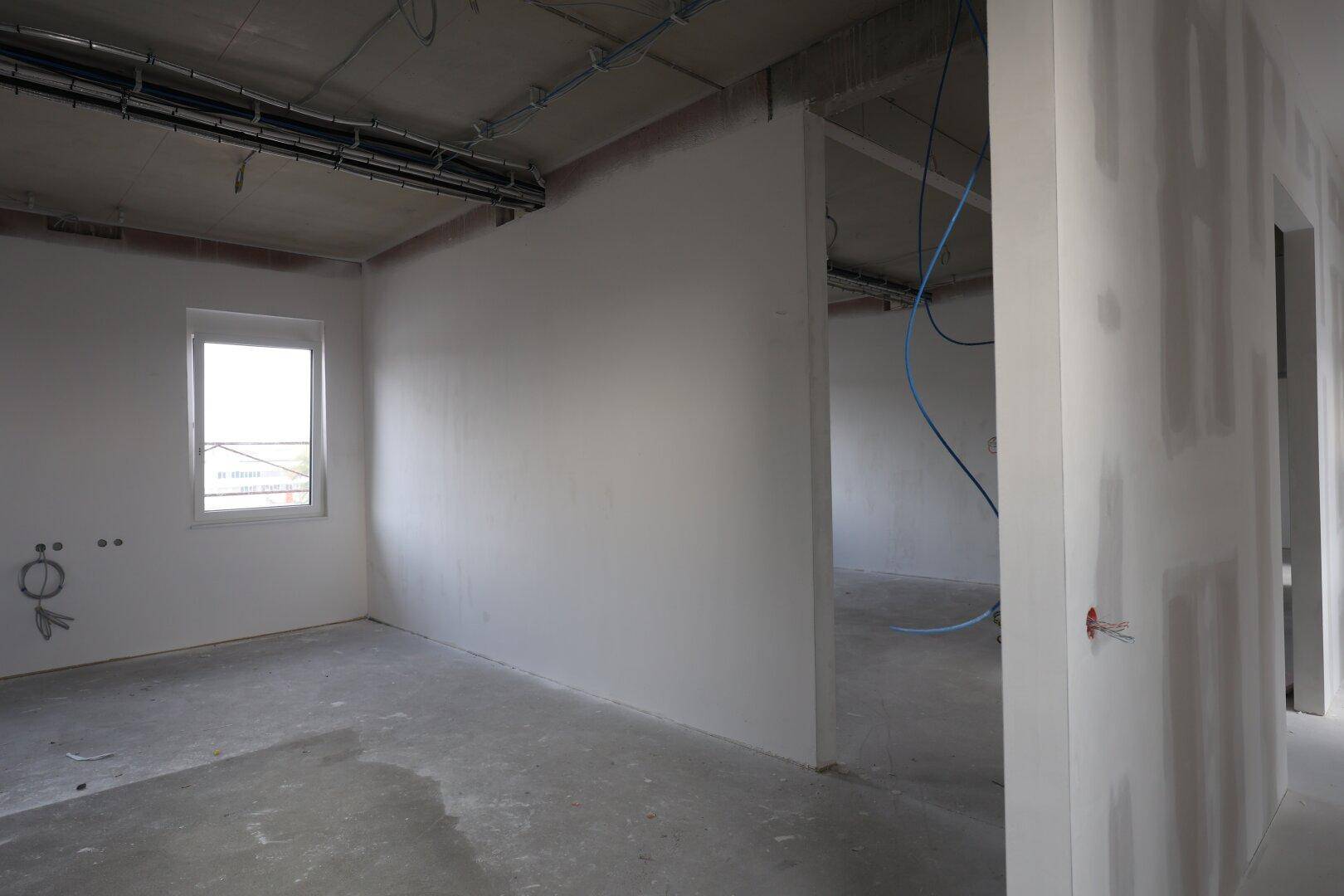 Unfertiger Innenraum mit Gipskartonwänden, Betonboden und sichtbaren Leitungen.