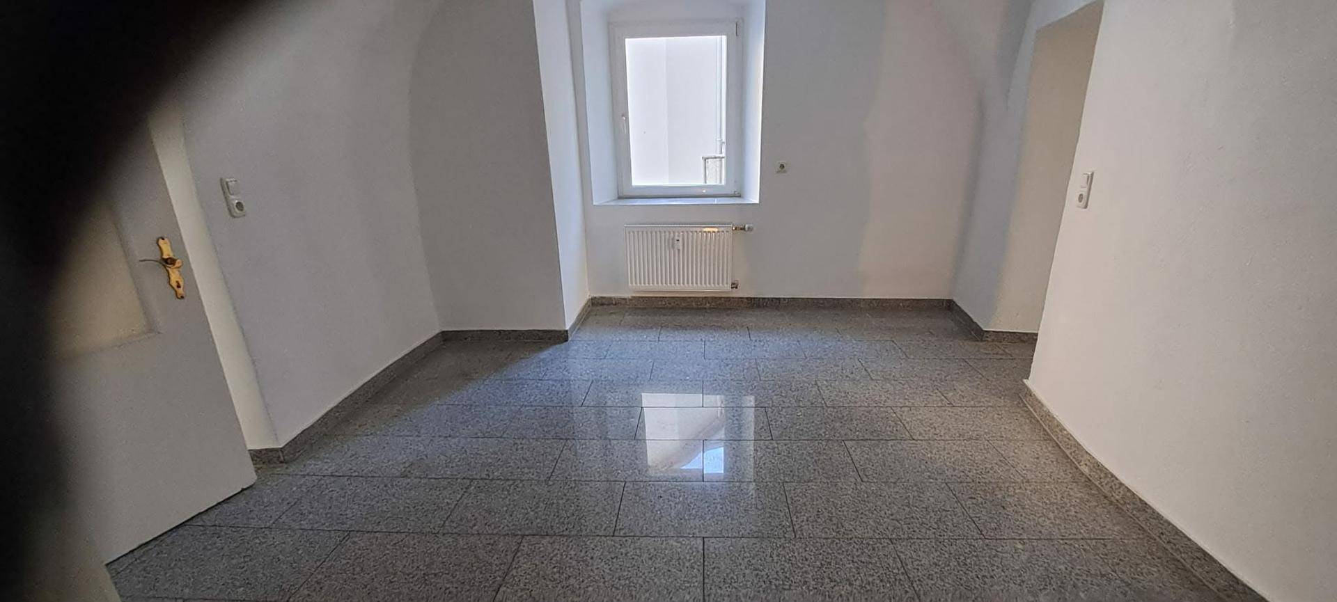 Leerer Raum mit grauen Fliesen und einem Fenster, das den Raum erhellt.