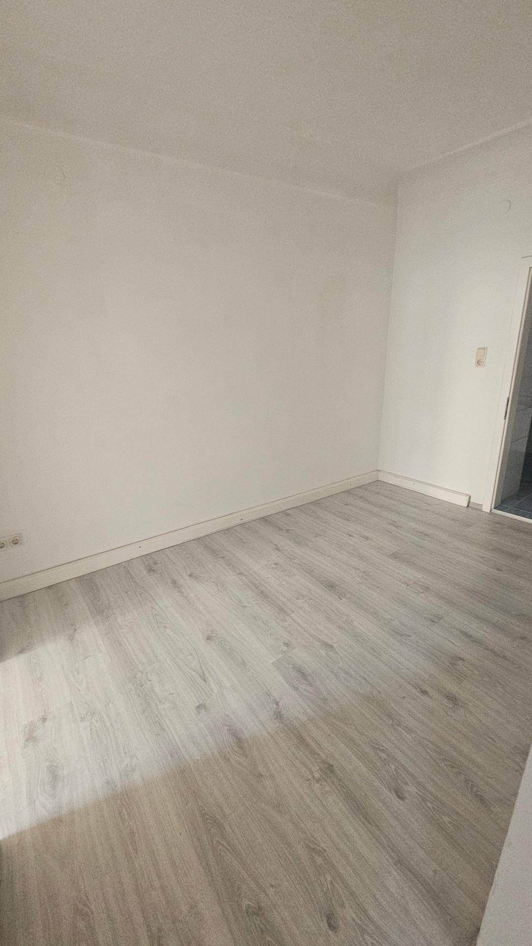 Einfaches, unmöbliertes Zimmer mit weißen Wänden und hellem Laminatboden.