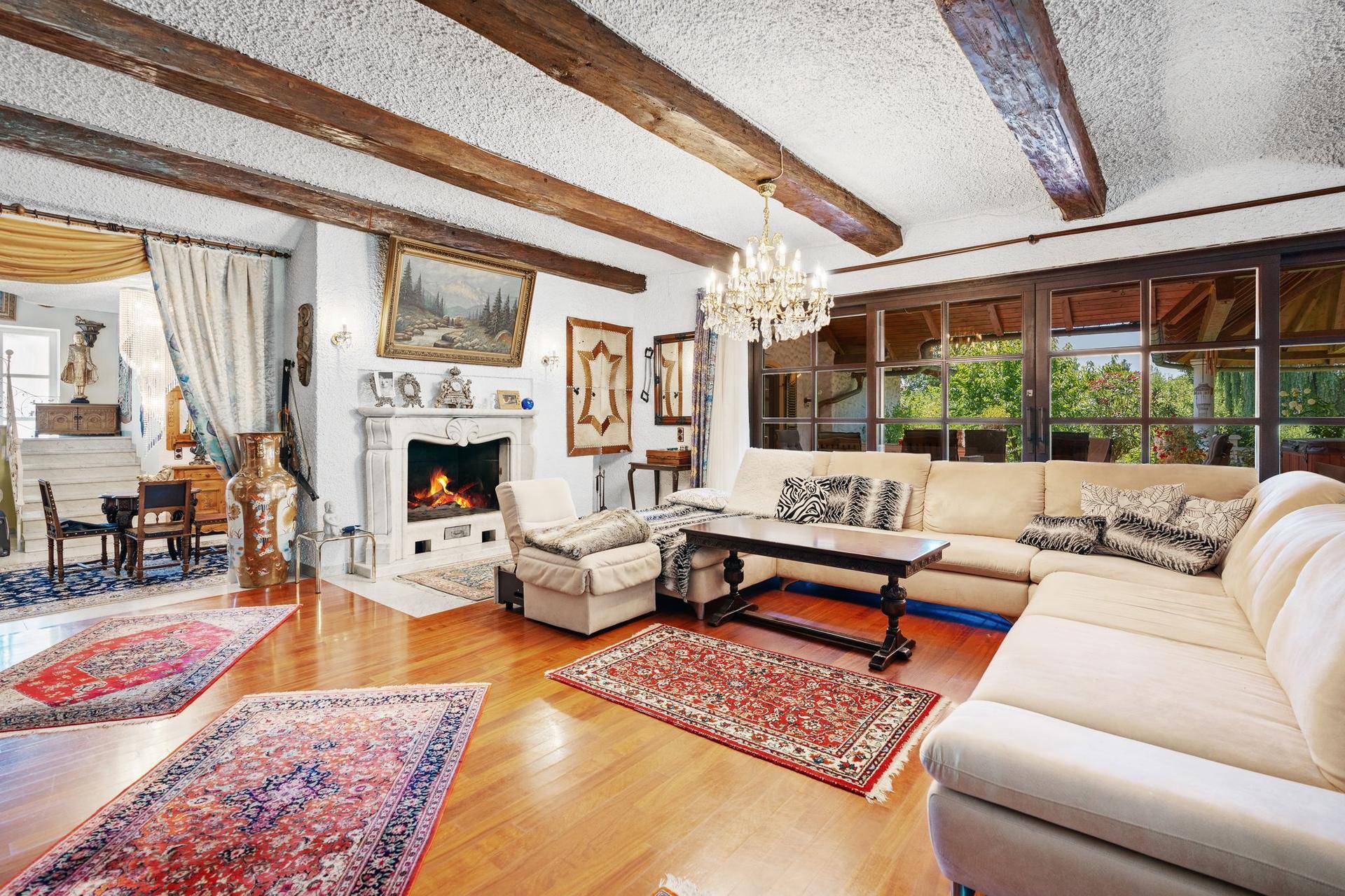 Elegantes Wohnzimmer mit Kamin, großem Sofa, Holzbalkendecke und Blick in den Garten.