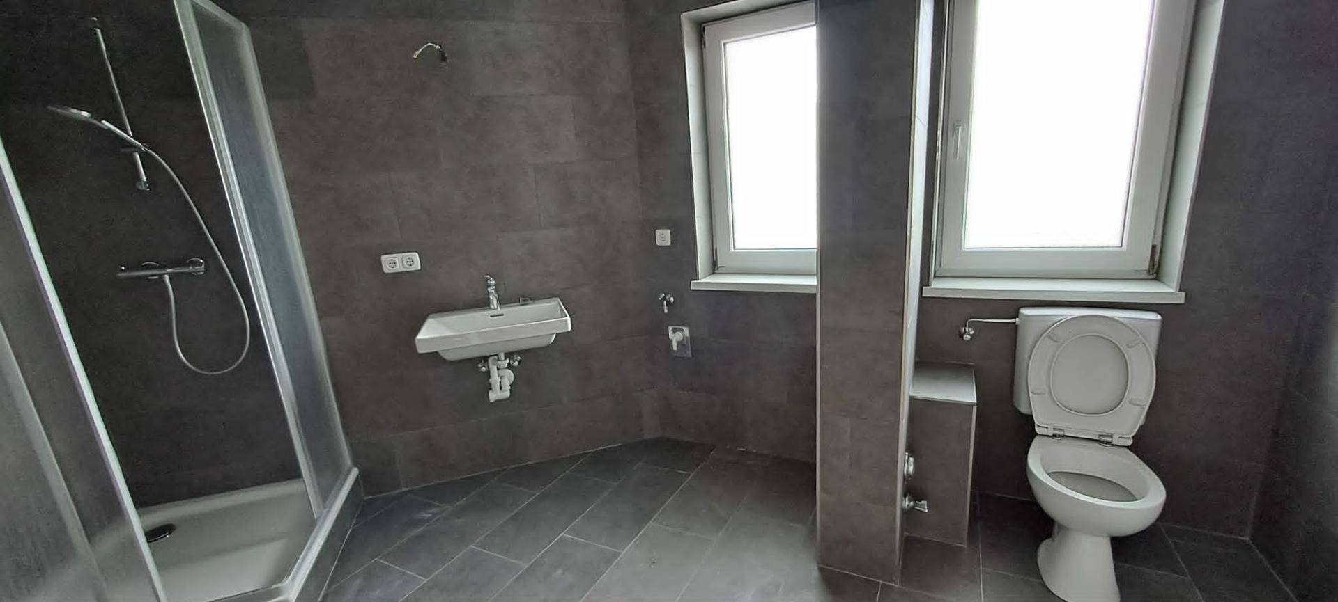 Modernes Badezimmer mit dunklen Fliesen, einer Duschkabine, Toilette und zwei Fenstern.