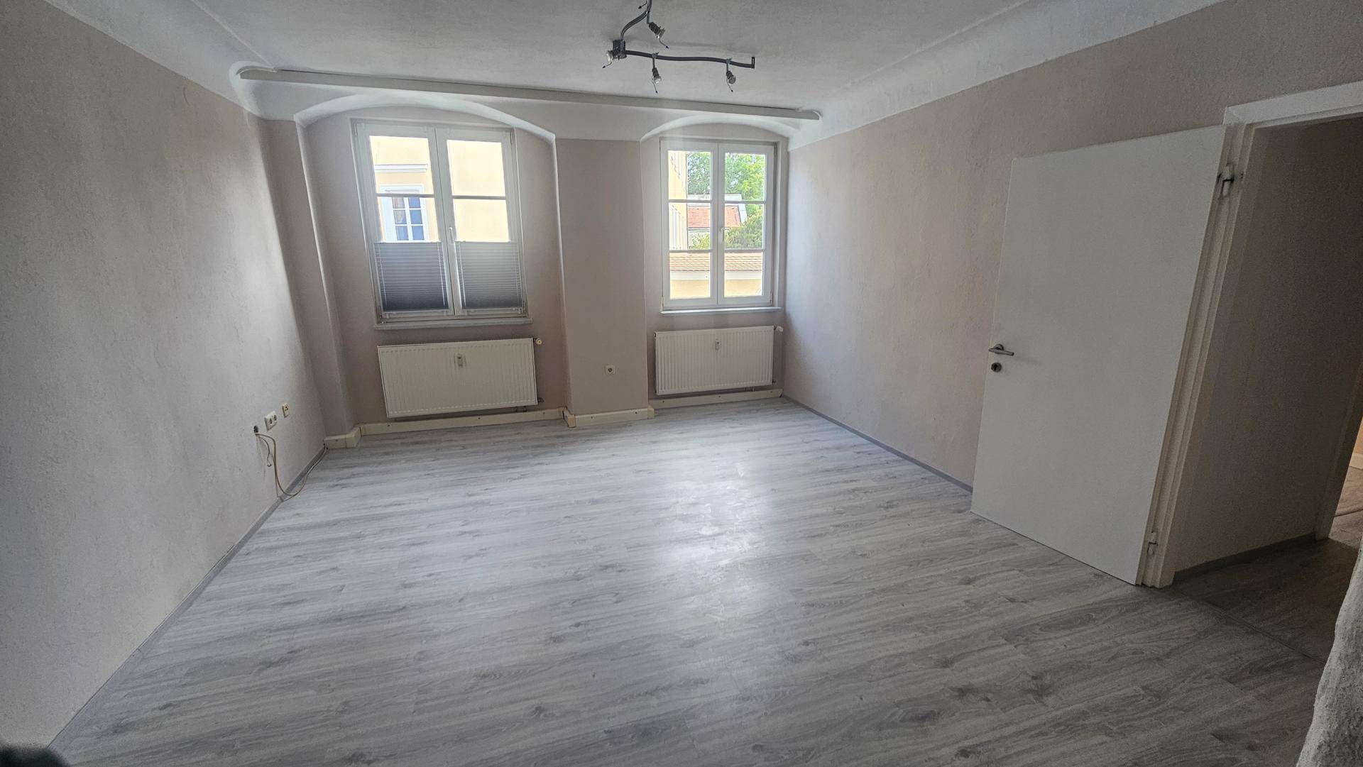 Renoviertes Zimmer mit hellem Laminatboden, zwei Fenstern und Zugang zu weiteren Räumen.