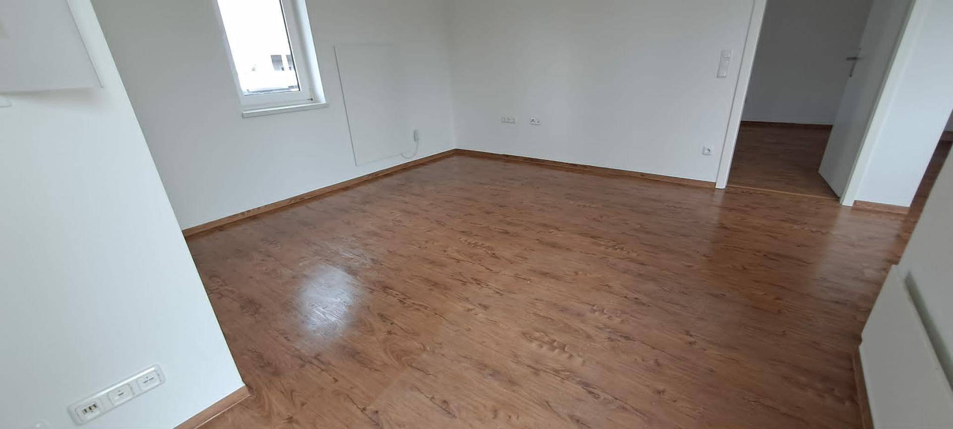 Leerer Raum mit Laminatboden, weißen Wänden und einem Fenster, das für natürliches Licht sorgt.