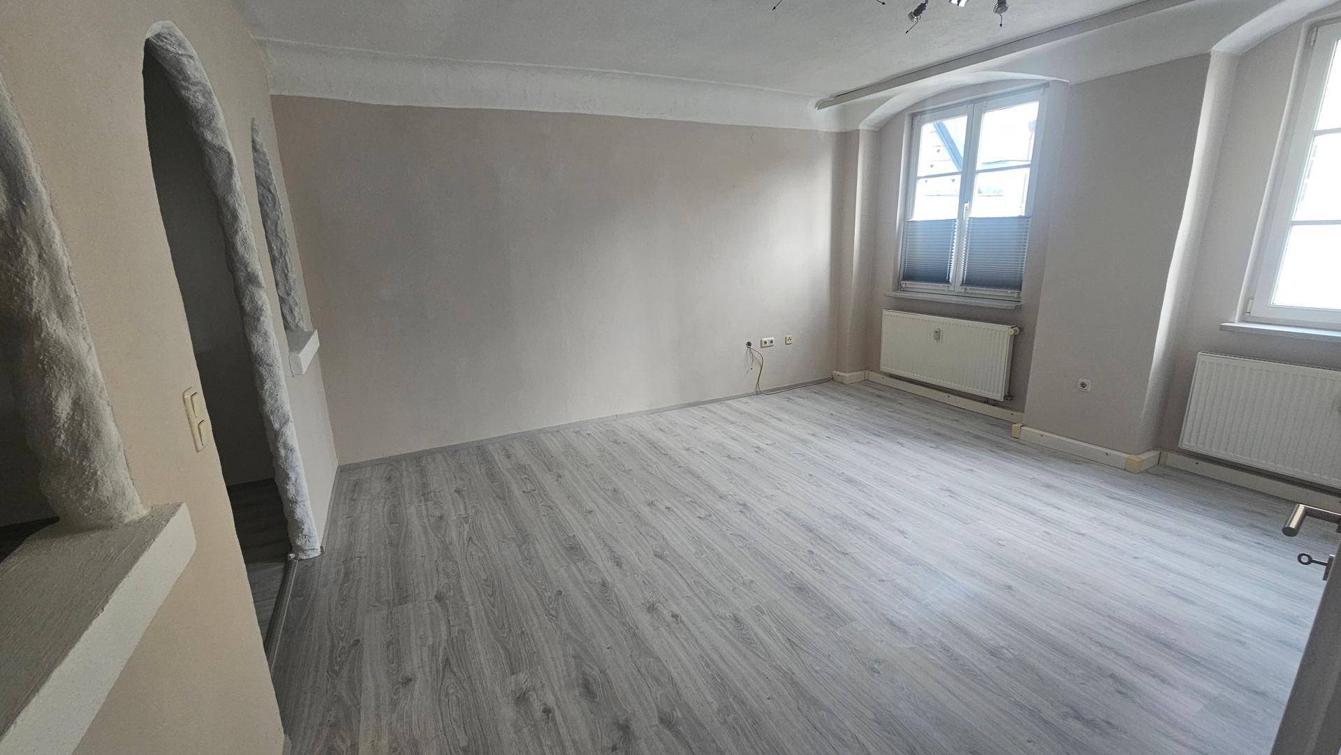 Helles Zimmer mit zwei Fenstern, Heizkörpern und neu verlegtem Laminatboden.