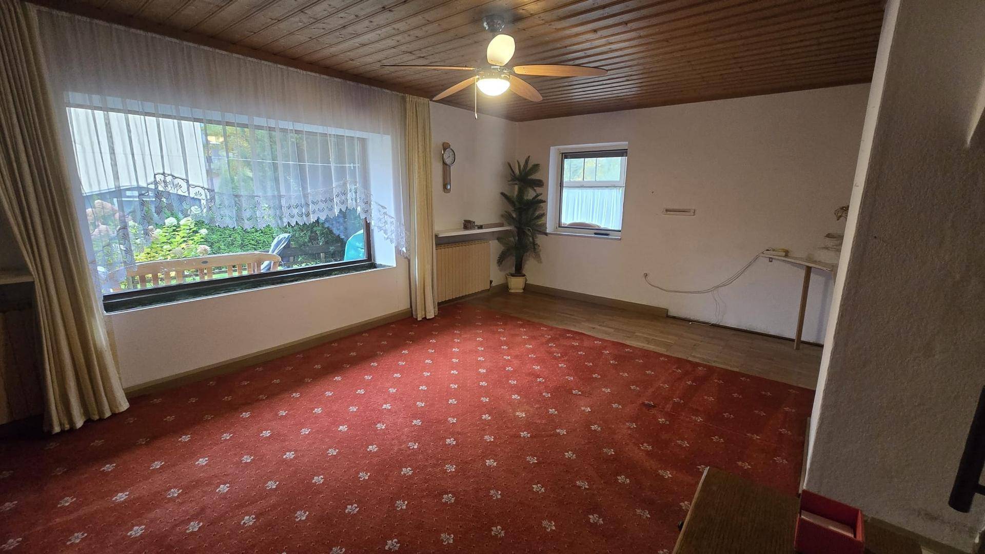 Geräumiges Wohnzimmer mit Holzdecke, rotem Teppich und einem großen Fenster mit Blick in den Garten.