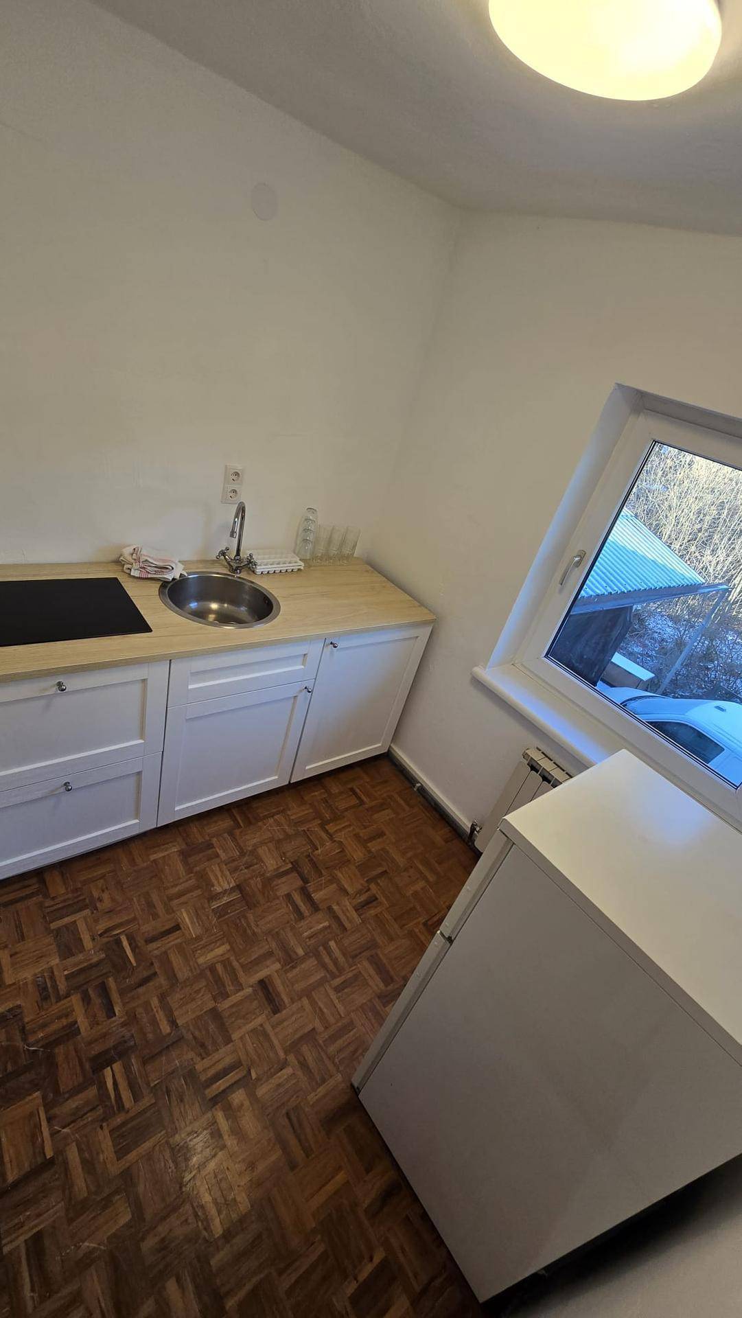 Kleine Küche mit Holzparkett, weißen Unterschränken und einem Fenster mit Blick nach draußen.