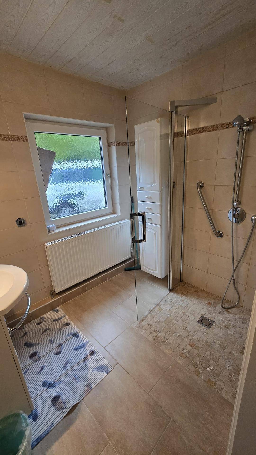 Helles Badezimmer mit beigen Fliesen, Duschkabine und einem Fenster für Tageslicht.