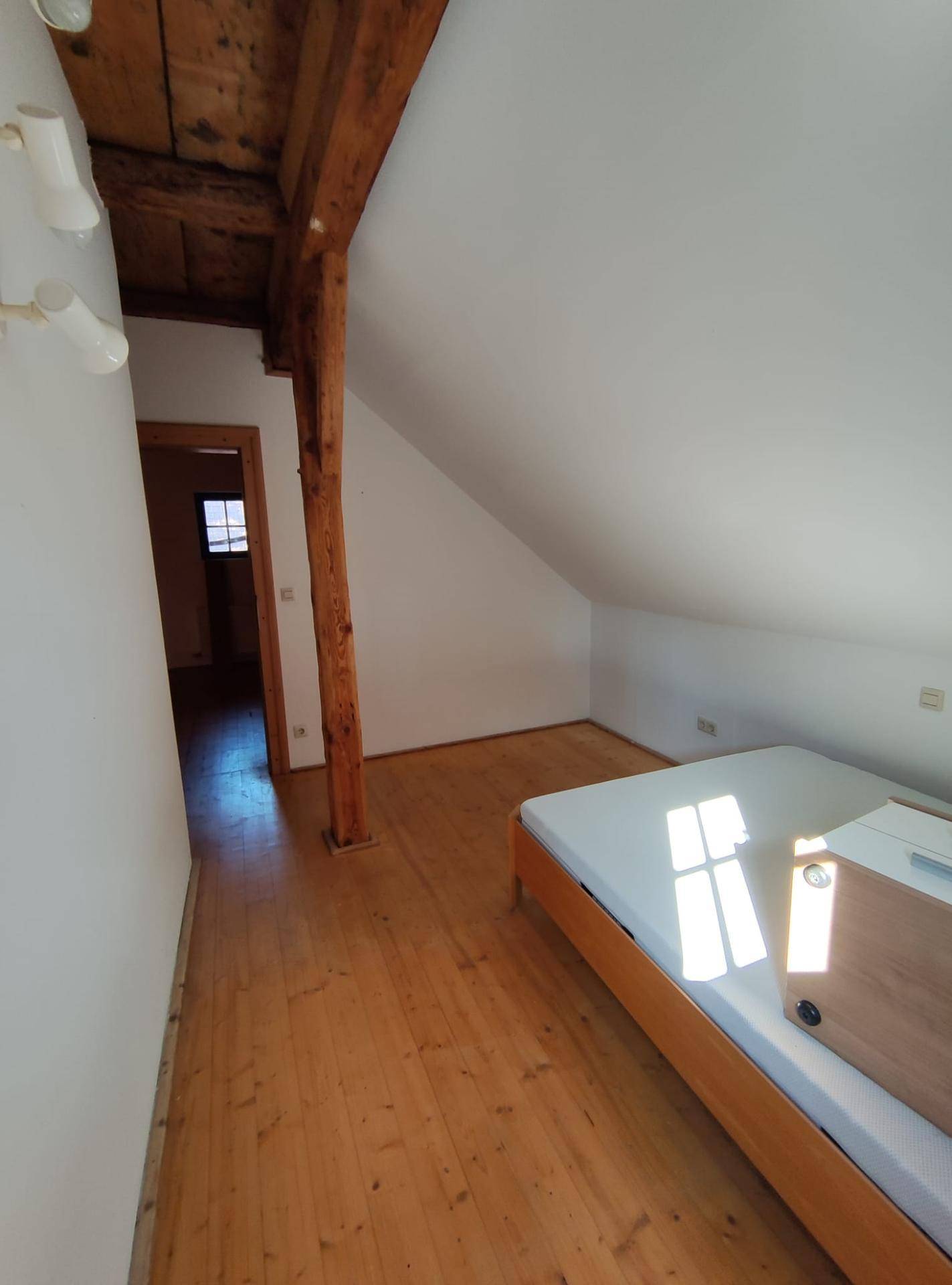 Geräumiges Schlafzimmer mit Holzboden, sichtbarem Holzbalken und Zugang zu einem weiteren Raum.
