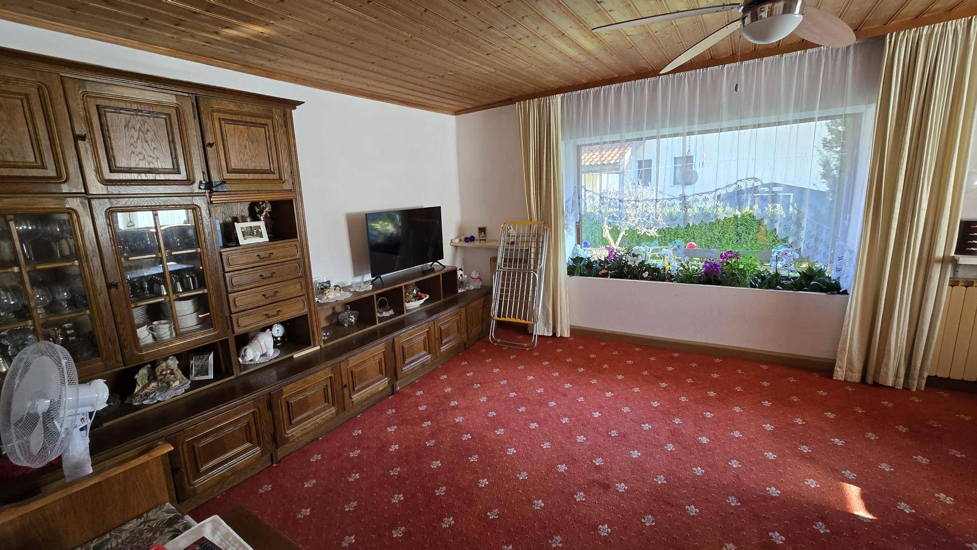 Wohnzimmer mit Holzdecke, rotem Teppich und einem großen Fenster mit Blick in den Garten.
