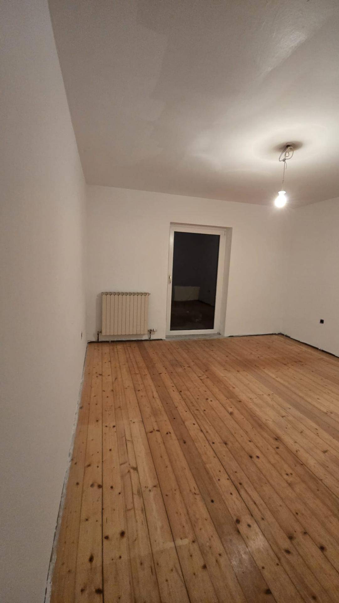 Helles Zimmer mit Holzdielenboden und Heizkörper, durch eine Tür gelangt man in einen weiteren Raum.