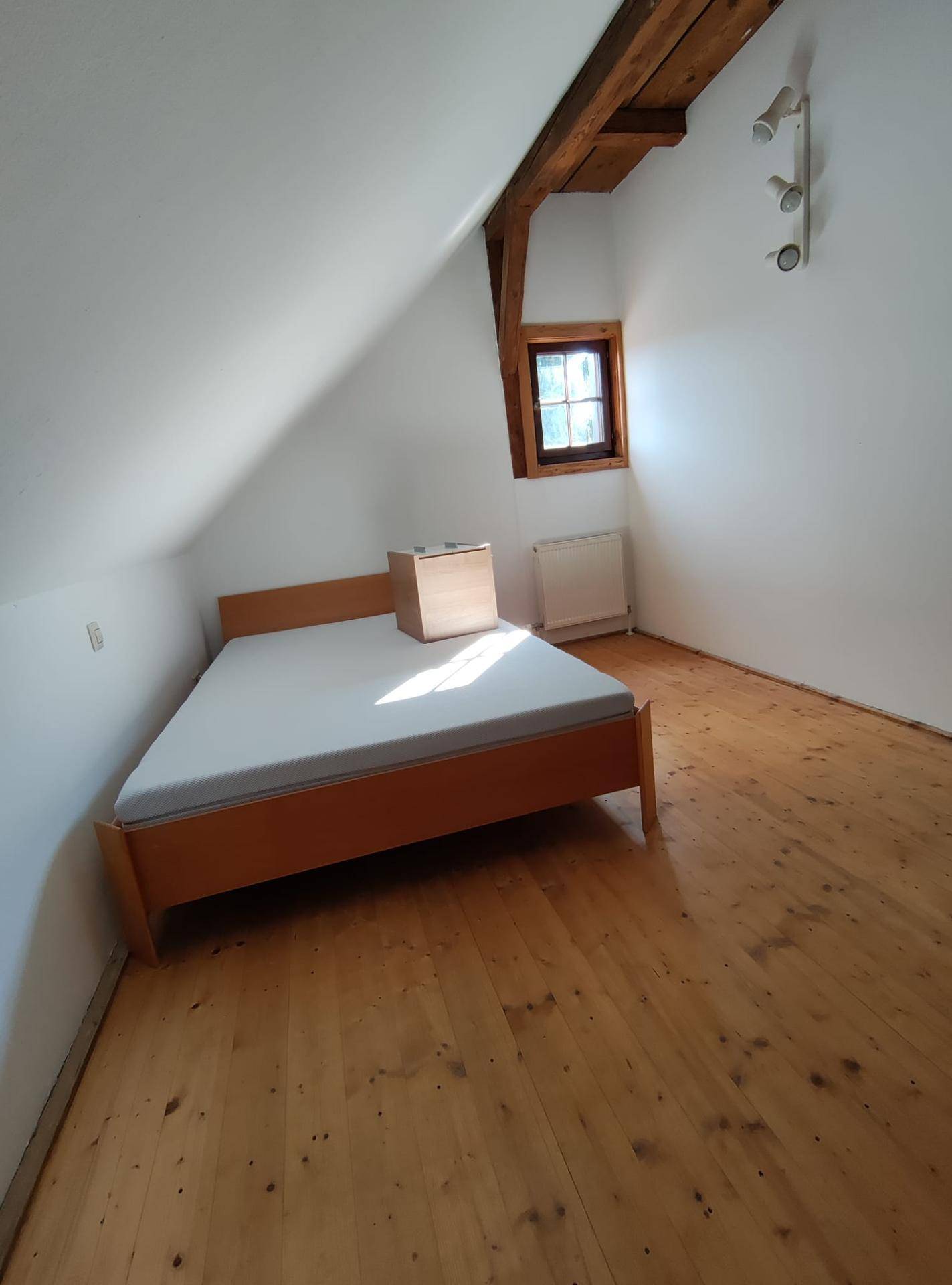 Helles Schlafzimmer unter dem Dach mit Holzboden, sichtbaren Balken und einem kleinen Fenster.