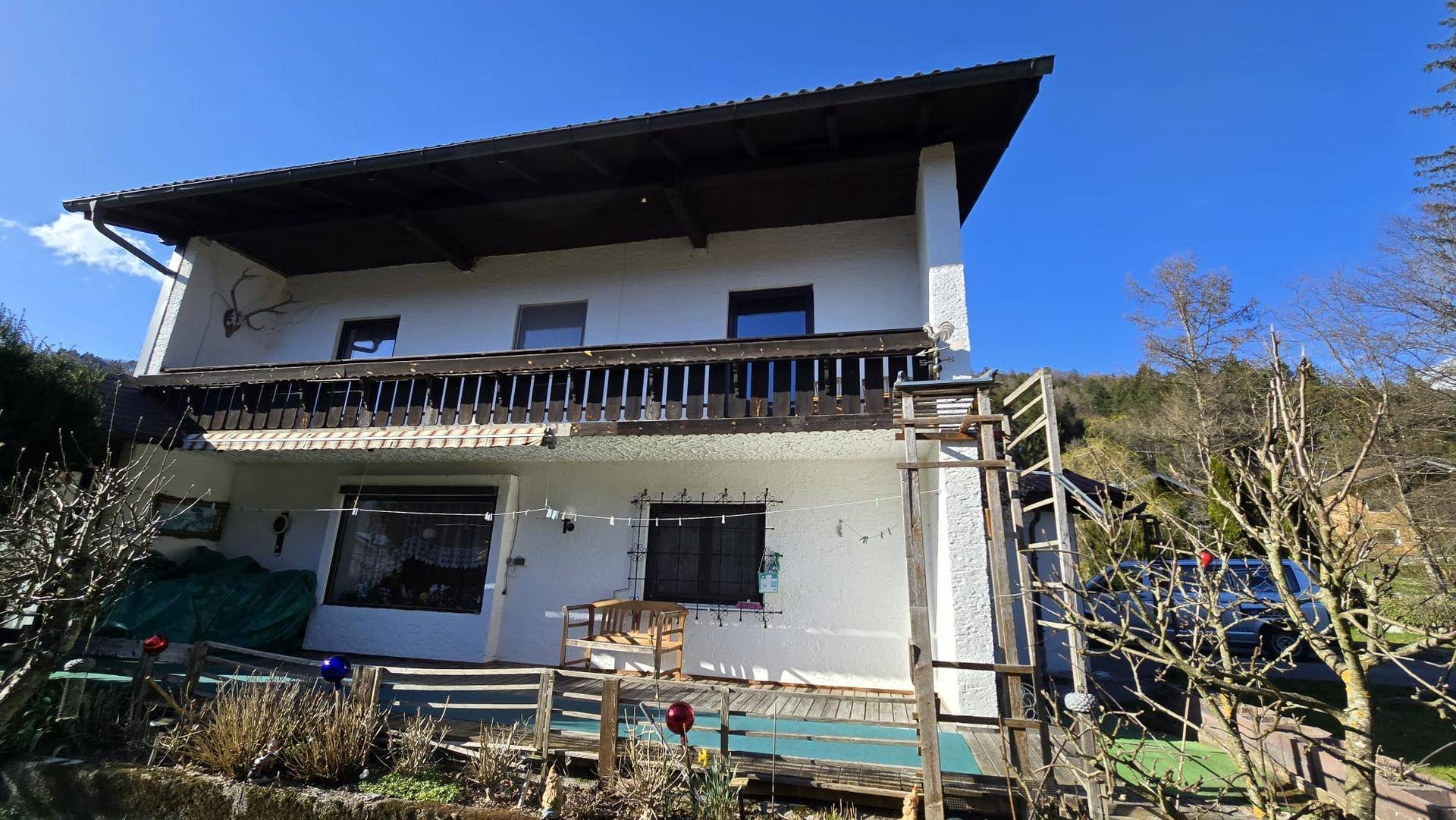 Weißes Haus mit traditionellem Holzbalkon und Blick auf die umliegende grüne Landschaft.