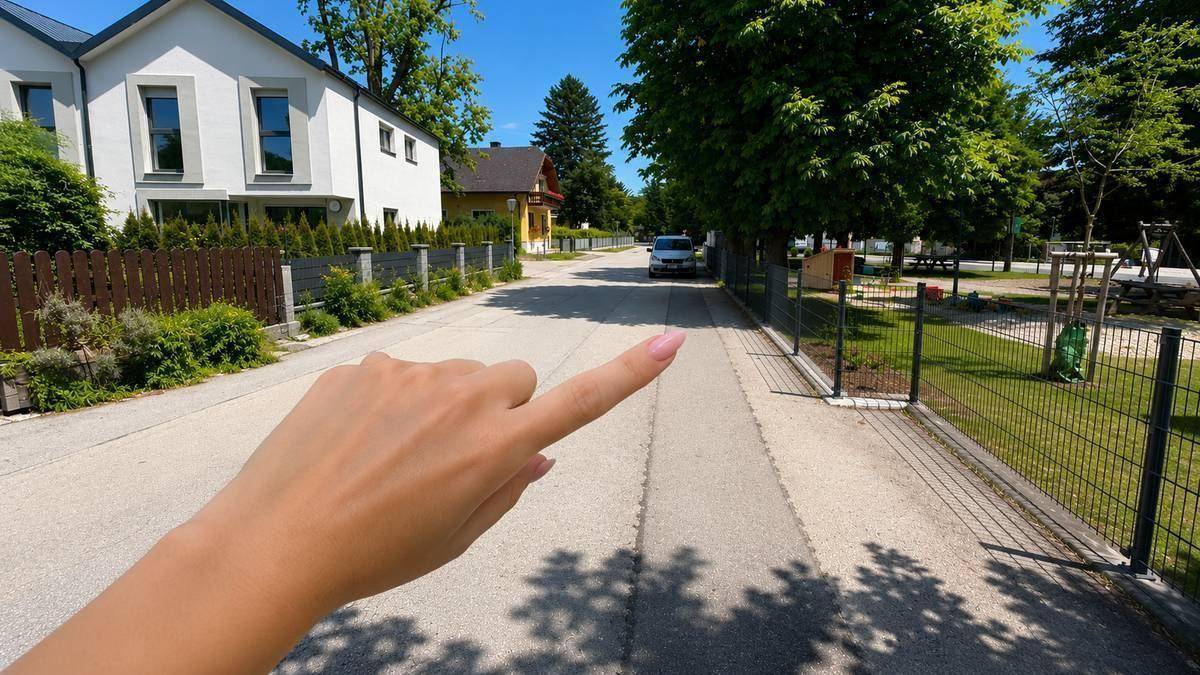 Eine Hand zeigt auf eine ruhige Wohnstraße mit modernen Häusern und einem grünen Zaun.