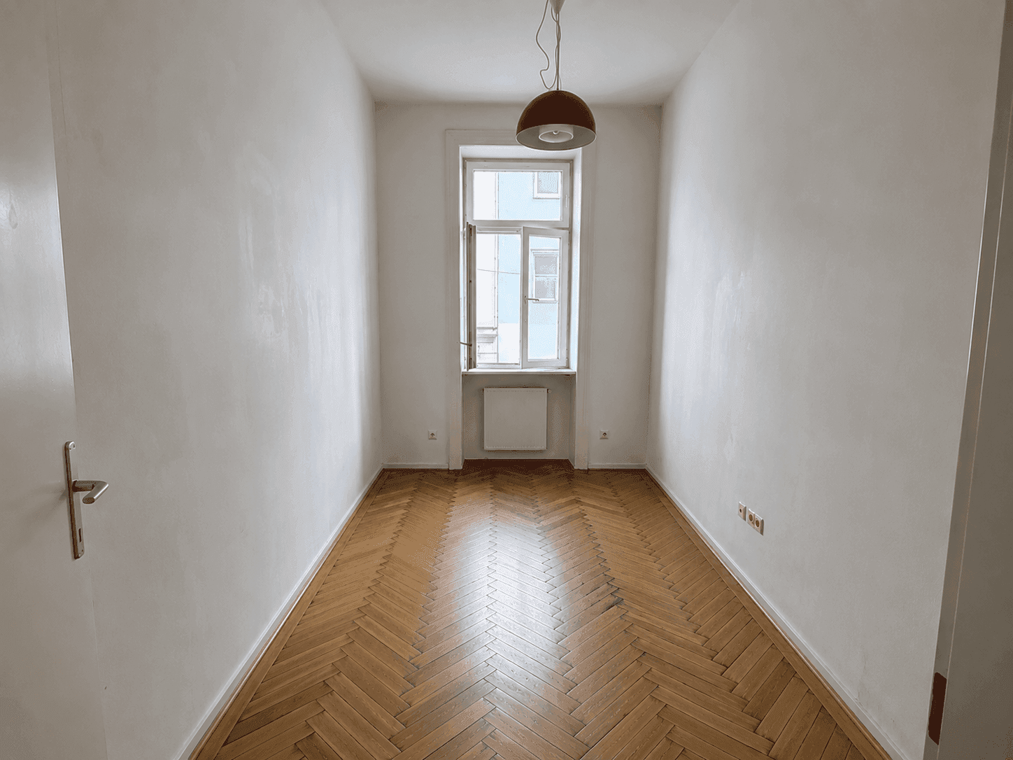Leerer Raum mit Fischgrätparkettboden, hohen Decken und einem großen Fenster, das viel Tageslicht hereinlässt.
