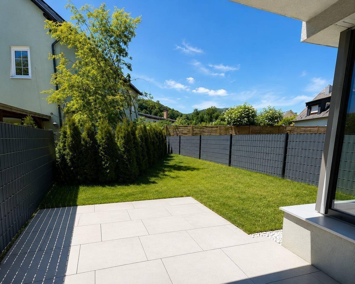 Privater Gartenbereich mit großer gepflasterter Terrasse und Rasenfläche, umgeben von einem Zaun.