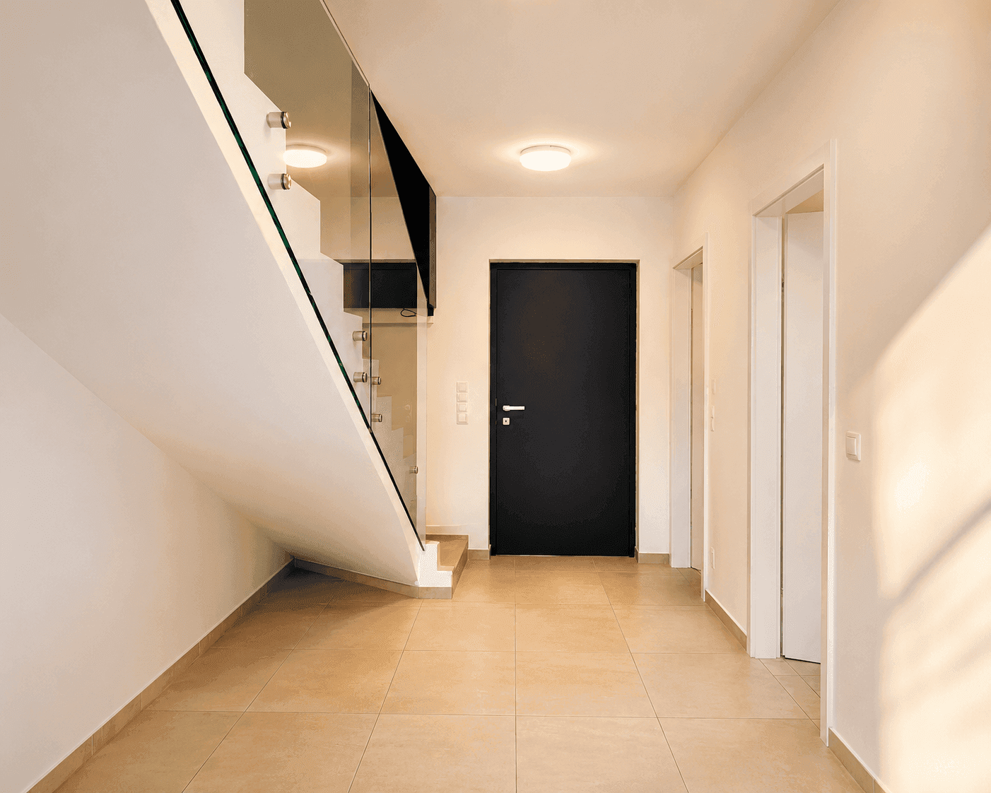 Heller Eingangsbereich mit modernen Fliesen, schwarzer Tür und eleganter Glasgeländer-Treppe.