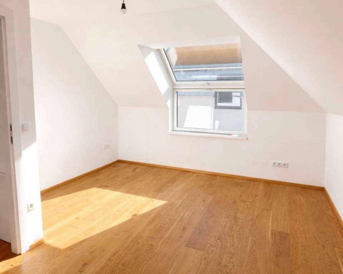Helles Dachgeschosszimmer mit Holzfußboden und schrägem Dach, das durch ein großes Fenster viel Licht erhält.