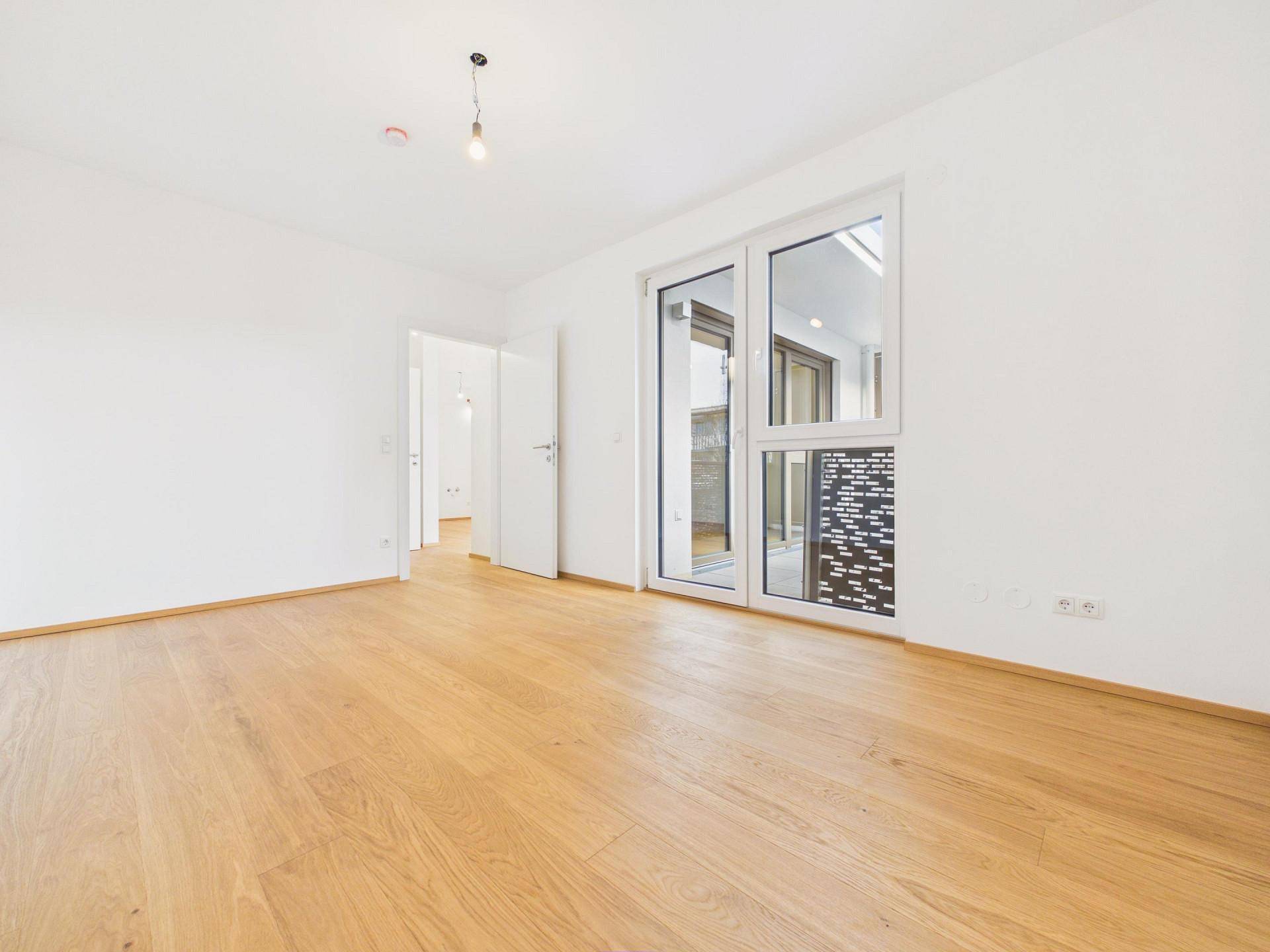 Leeres Schlafzimmer mit Holzboden, weiß gestrichenen Wänden und Zugang zu einem Außenbereich.