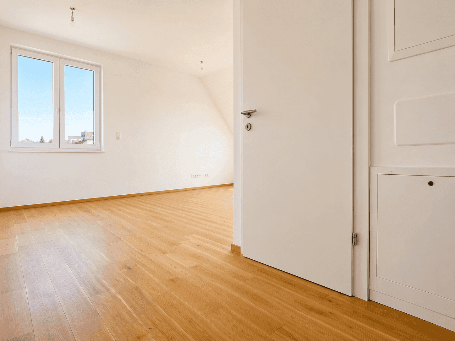 Leerer Raum im Dachgeschoss mit Holzboden, einem Fenster und einer weißen Tür.