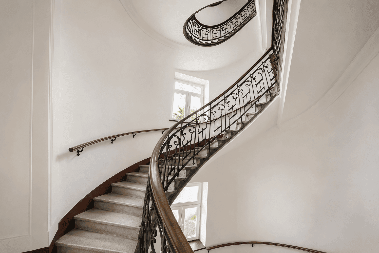 Elegantes Treppenhaus mit geschwungener Steintreppe, schmiedeeisernem Geländer und großen Fenstern für viel Licht.