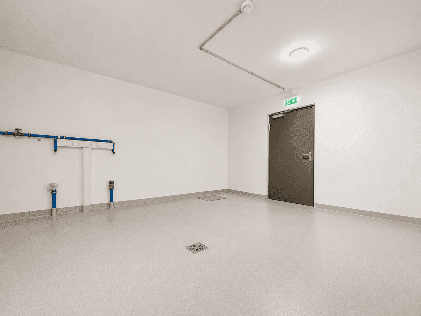 Leerer Abstellraum mit hellgrauem Boden und sichtbaren Rohrleitungen an der Wand.