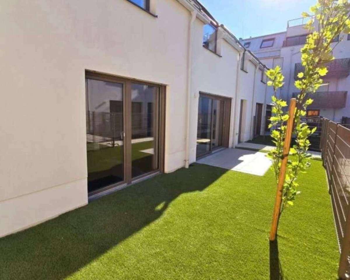 Kleiner privater Gartenbereich mit Kunstrasen und einem jungen Baum vor der modernen Gebäudefassade.