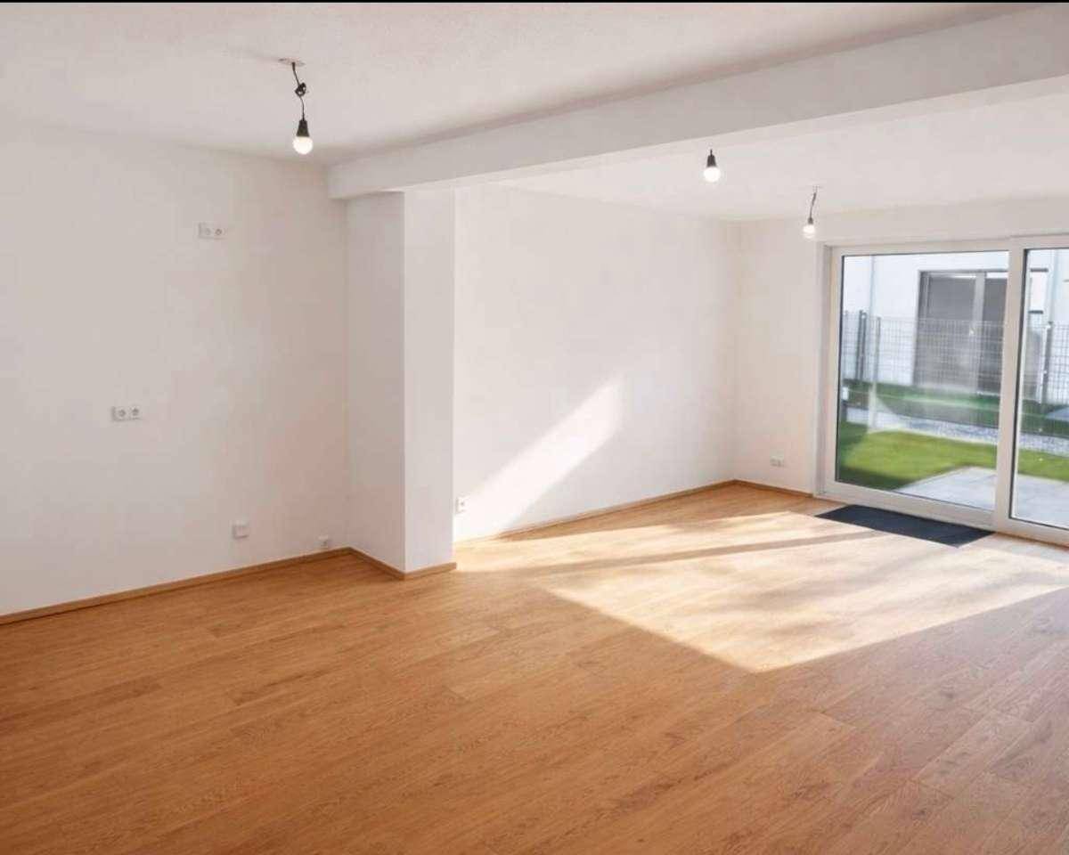 Geräumiges, leeres Wohnzimmer mit hellem Holzfußboden und großen Fenstern, die viel Tageslicht hereinlassen.