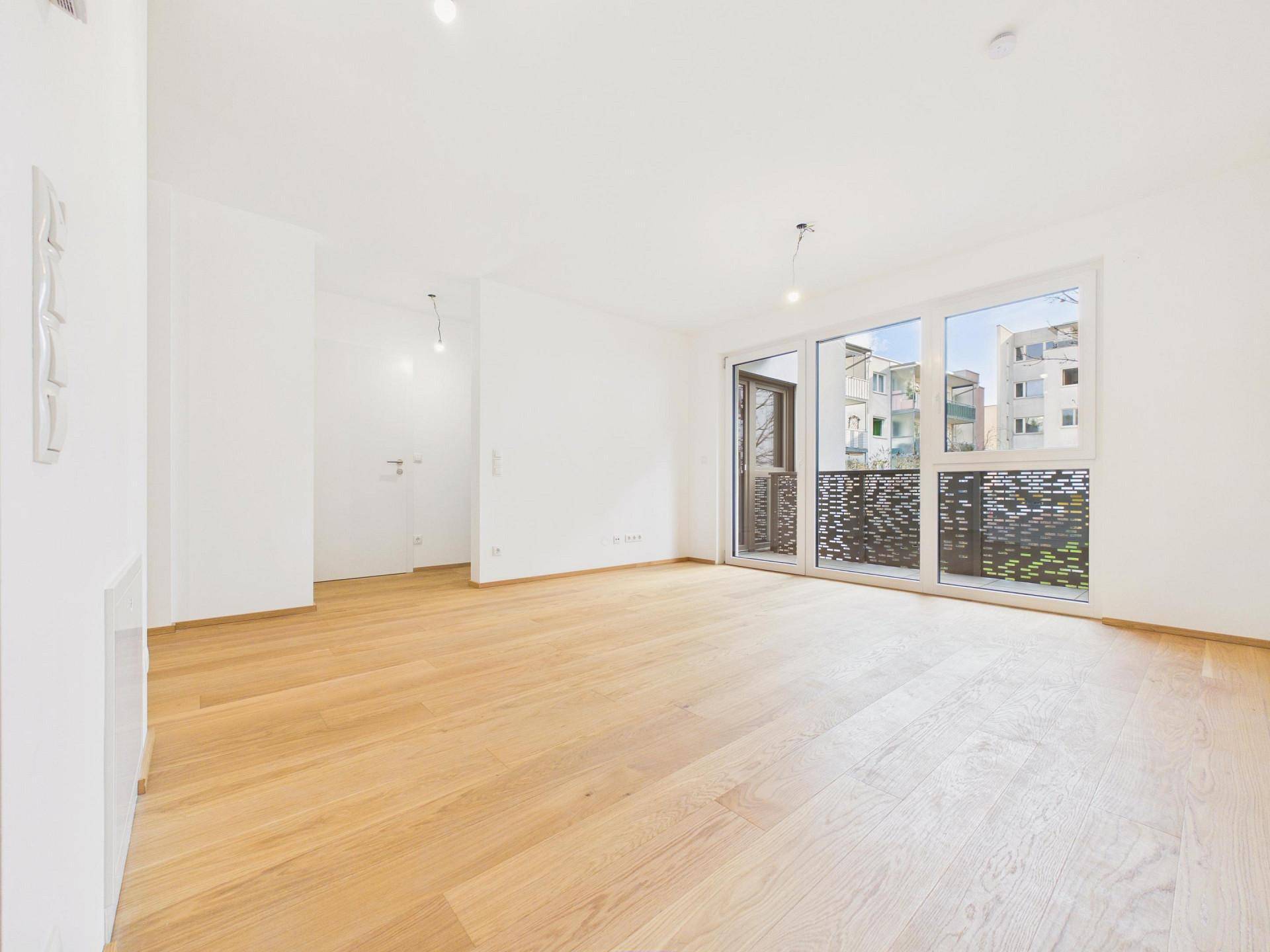 Helles, leeres Wohnzimmer mit Holzboden und großen Fenstern, die auf einen Balkon führen.