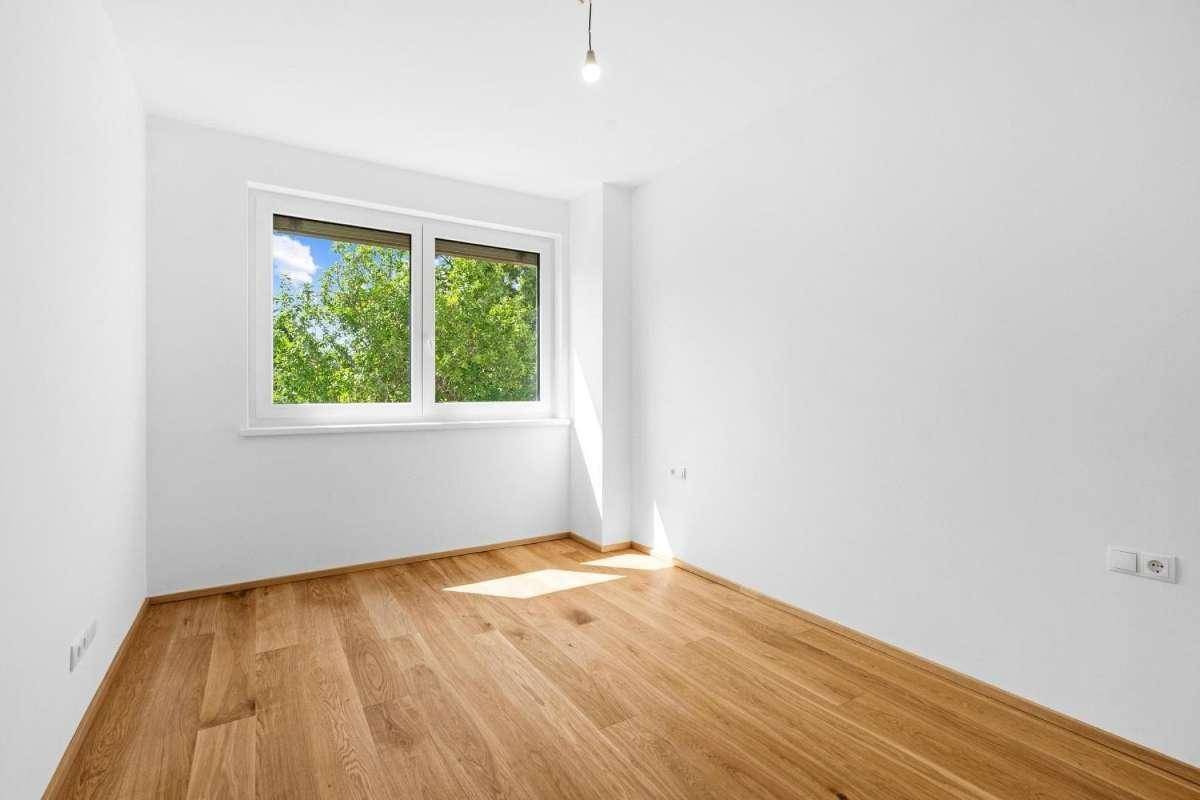 Leerer Raum mit Holzboden und großem Fenster, das viel Tageslicht hereinlässt.