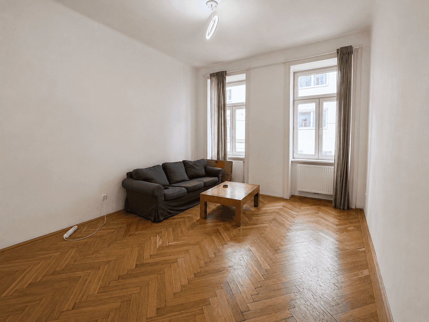 Gemütliches Wohnzimmer mit Fischgrätparkett, Sofa und zwei großen Fenstern.