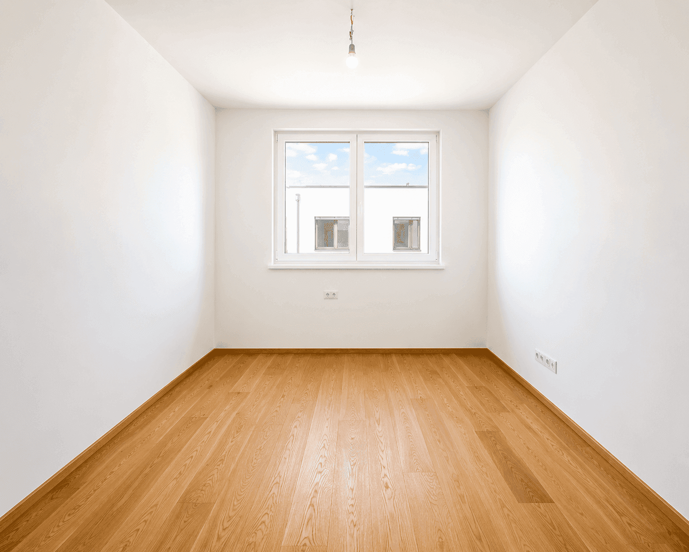 Leerer Raum mit Holzboden und Fenster, das auf ein gegenüberliegendes Gebäude blickt.