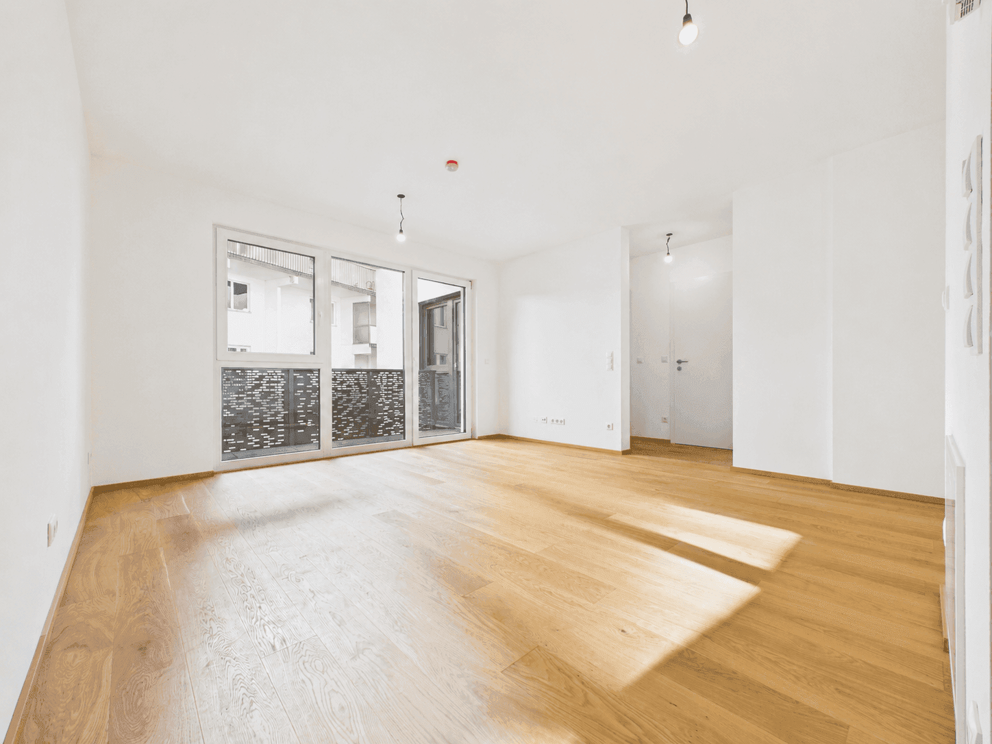 Helles, leeres Wohnzimmer mit großen Fenstern, Holzboden und Zugang zum Balkon.