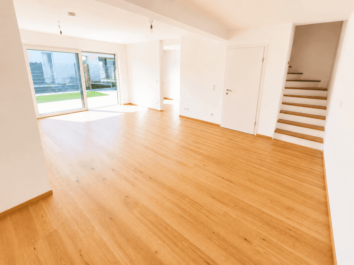 Geräumiges, leeres Wohnzimmer mit hellem Holzfußboden, weißen Wänden und Zugang zu einer Terrasse.