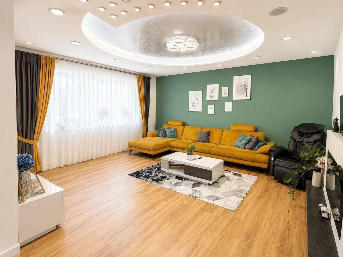 Geräumiges Wohnzimmer mit gelbem Ecksofa, grün akzentuierter Wand und moderner Deckenbeleuchtung.