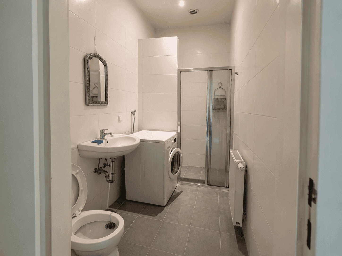 Funktionales Badezimmer mit Dusche, WC, Waschbecken und Waschmaschine in hellen Fliesen.
