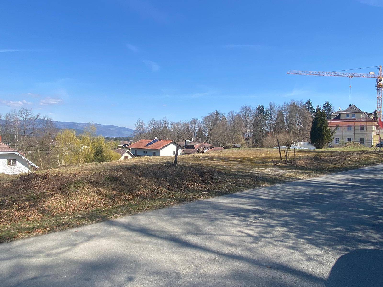 Blick über ein offenes Grundstück mit Straße im Vordergrund und Baustelle mit Kran in der Ferne.