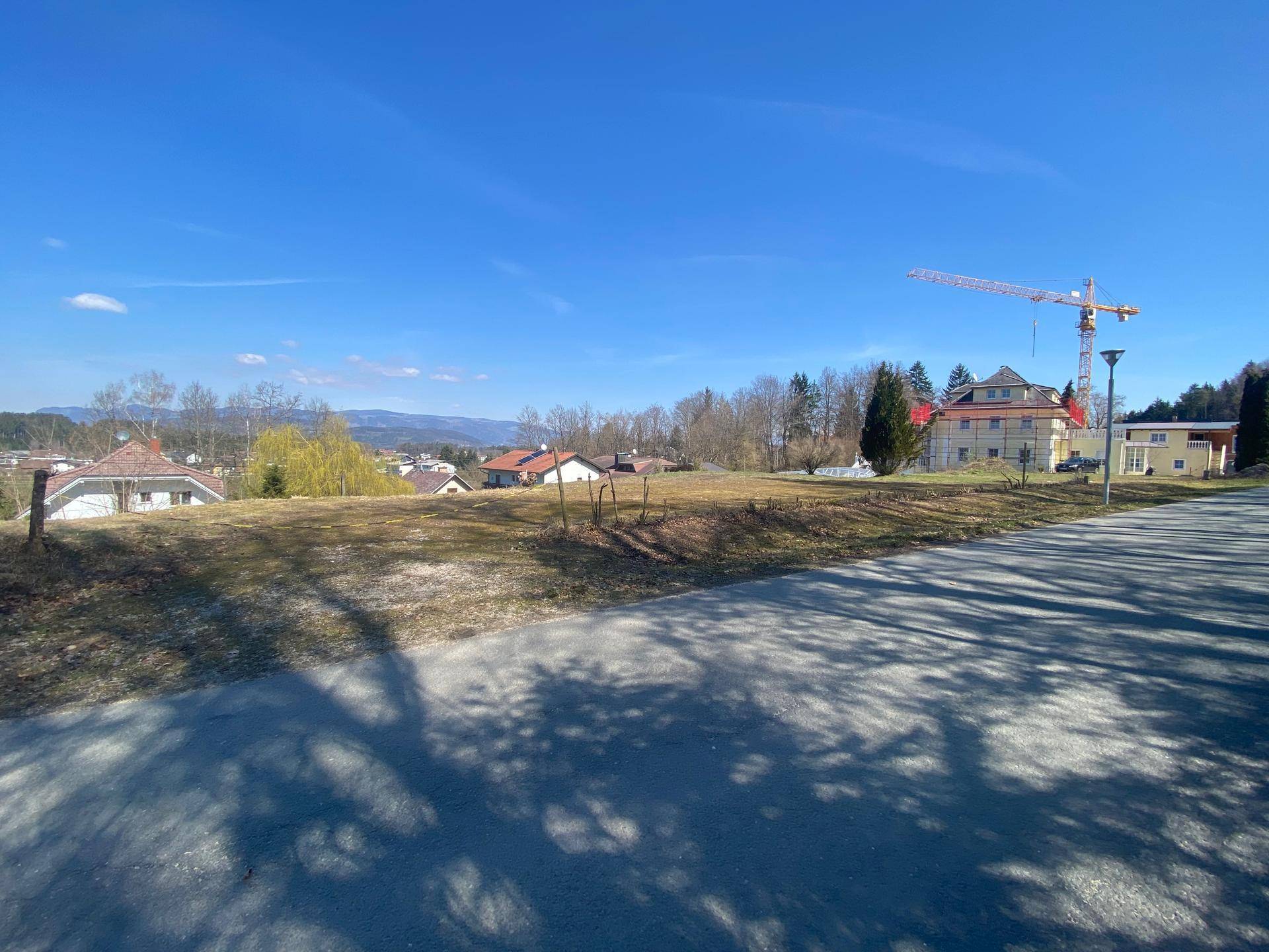 Blick über ein offenes Grundstück mit Straße im Vordergrund und Baustelle mit Kran in der Ferne.