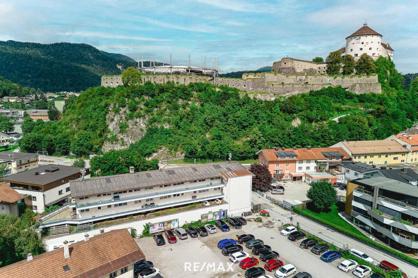 Luftaufnahme eines Gebäudes mit Parkplatz, umgeben von grünen Hügeln und einer historischen Festung im Hintergrund.