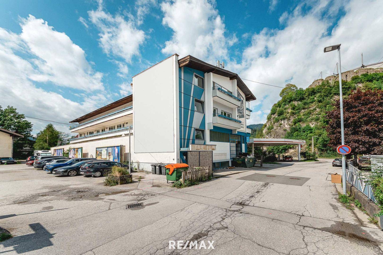 Ansicht eines modernen Geschäftsgebäudes mit blauen Akzenten, Parkplätzen und einer Bergkulisse im Hintergrund.