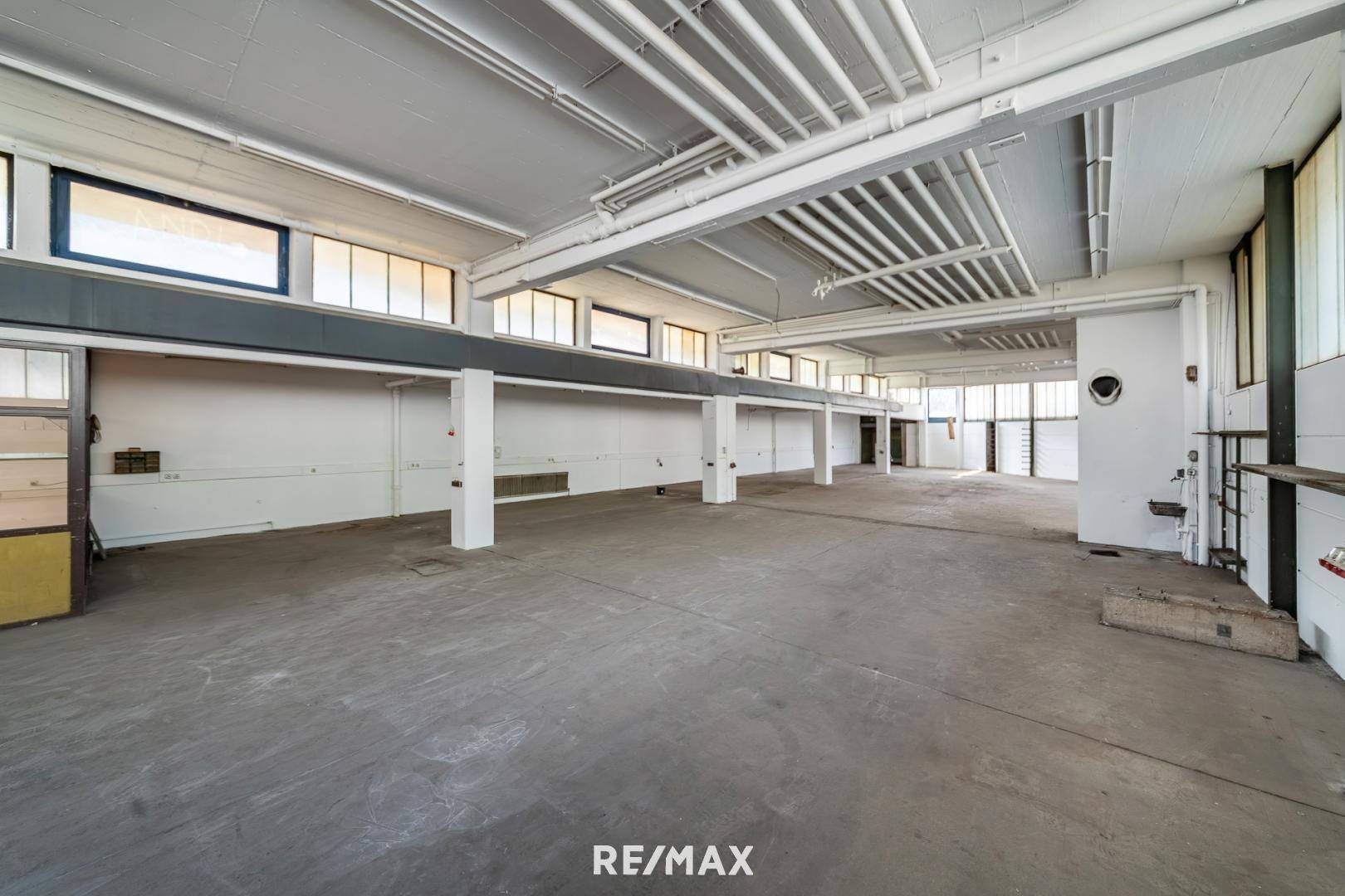 Weitläufiger, leerer Gewerberaum mit Betonboden, sichtbaren Rohren an der Decke und großen Fenstern.