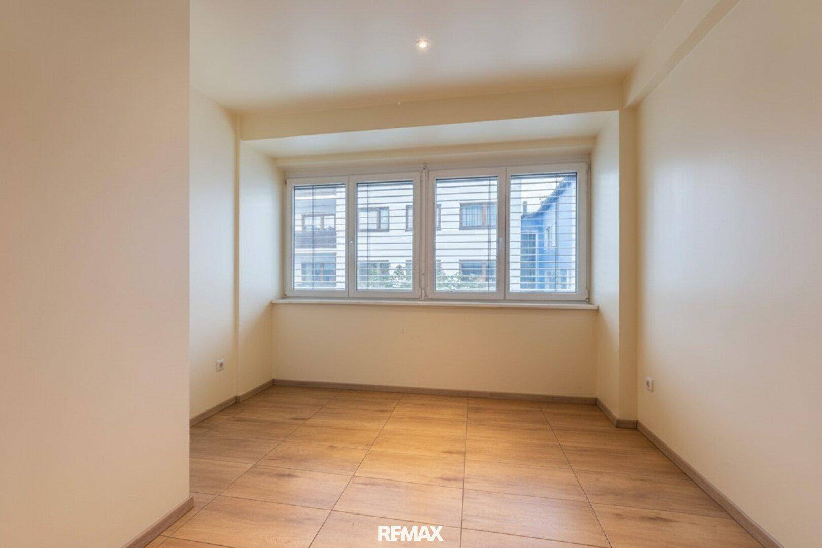 Helles Zimmer mit großen Fenstern und Jalousien, gefliestem Boden und neutralen Wänden.